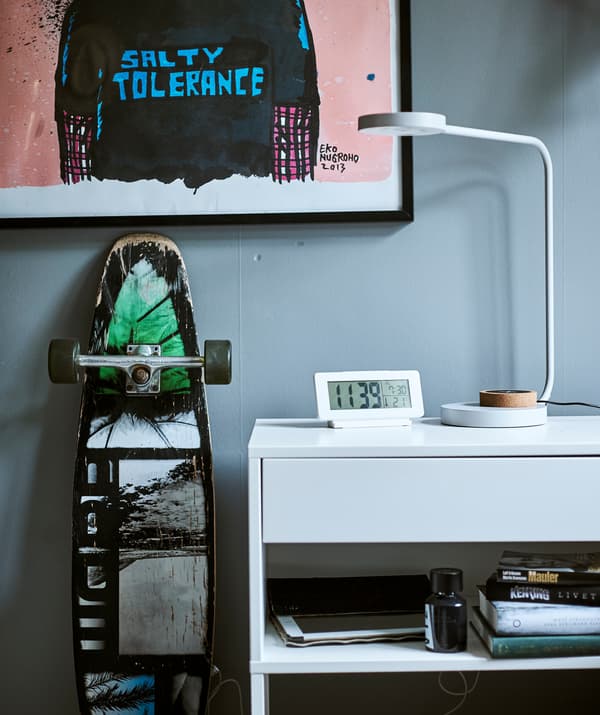 Un bureau blanc avec une tablette encastrée, au-dessus se trouve une lampe de bureau et une horloge digitale, un cadre avec un graffiti est suspendu au mur au-dessus d’un skateboard.