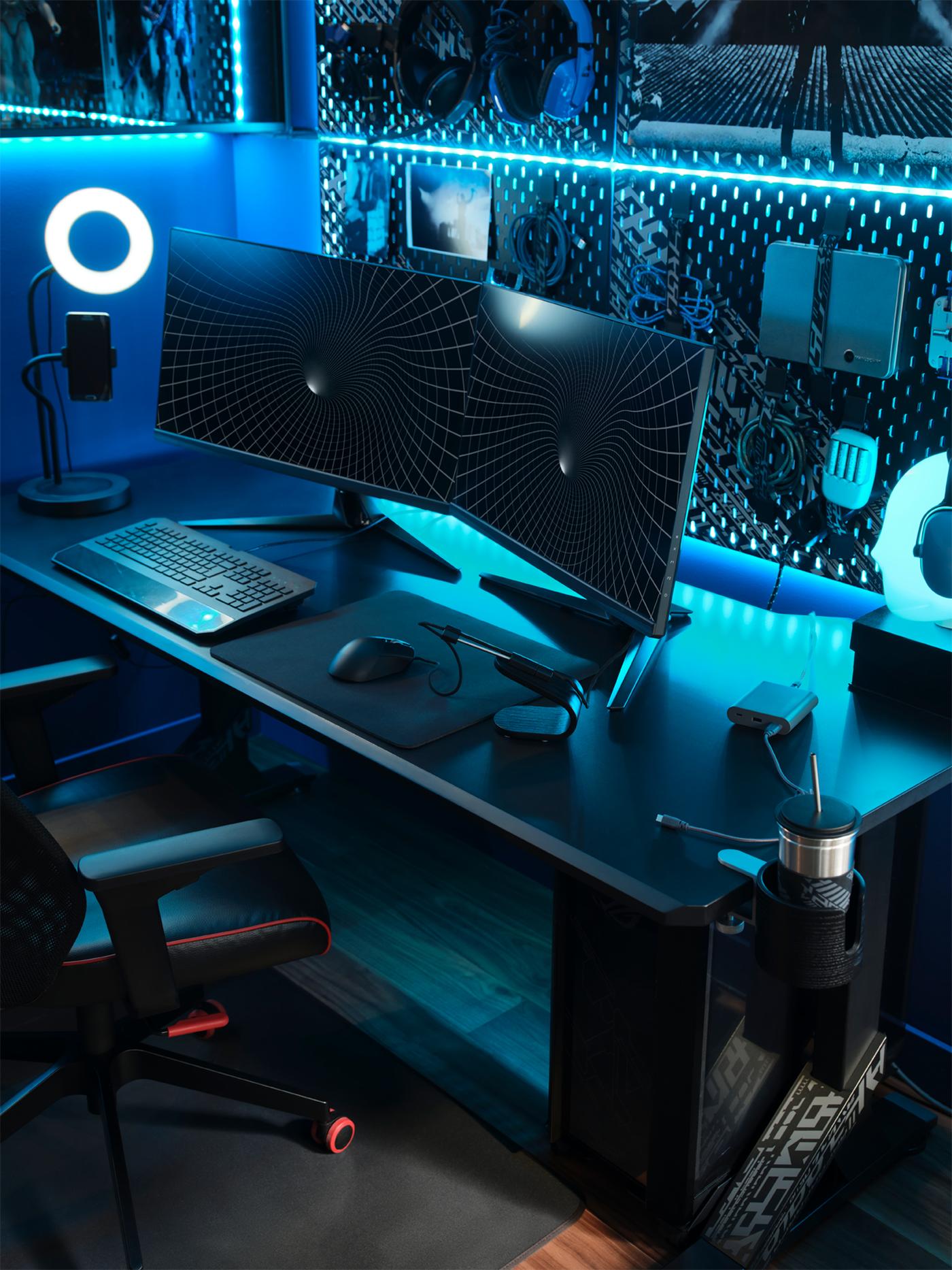 Un bureau de jeu UPPSPEL noir sur lequel se trouvent deux écrans, un anneau lumineux et un clavier, devant un panneau perforé sur lequel sont posés des accessoires technologiques.