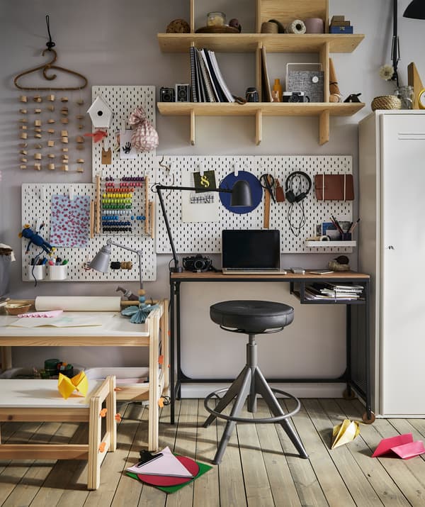 Un bureau d’enfant avec du matériel de bricolage à côté d’un bureau d’adulte avec un tabouret et des panneaux perforés sur le mur.