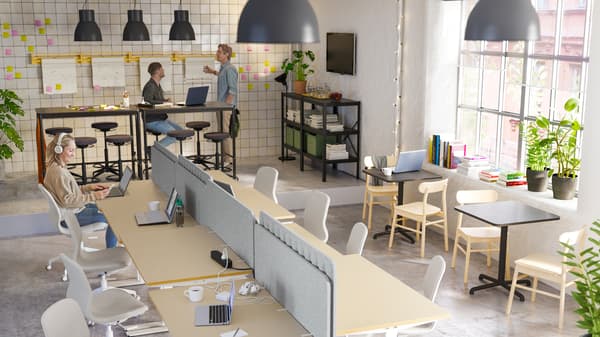 Un bureau ouvert où deux hommes travaillent à des tables hautes, et une femme rédige des e-mails assise sur une chaise beige/blanche à un bureau.
