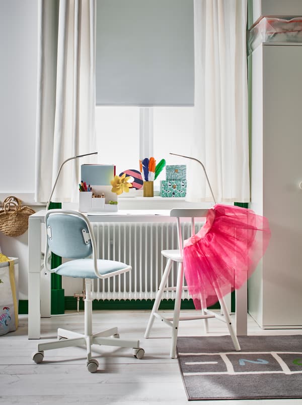 Un bureau PÅHL blanc et une chaise junior AGAM à côté d’une fenêtre ensoleillée, dans une chambre d’enfant claire au sol en bois.