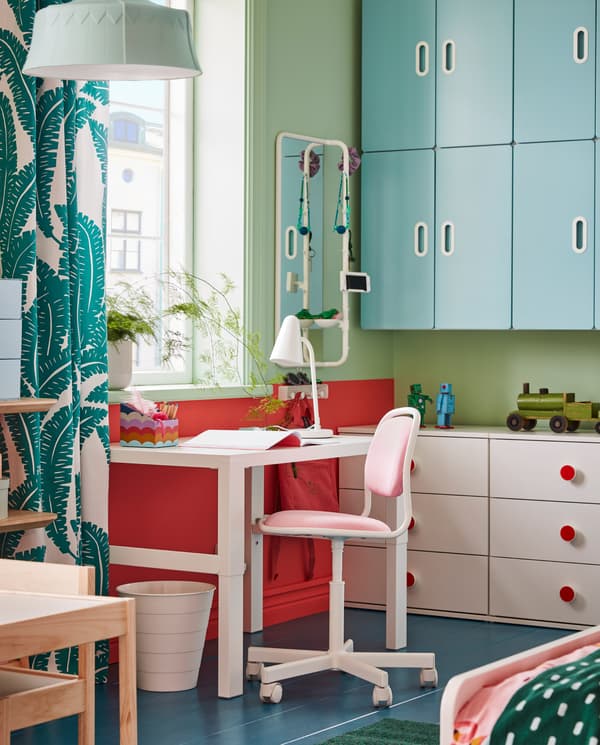 Un bureau PÅHL, une chaise de bureau pour enfants rose/blanc, une corbeille blanche et une lampe de table blanche, près d’une fenêtre.