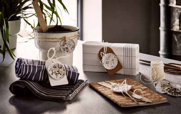 Un cache-pot beige, un carnet brun, des serviettes en tissu et en papier avec des dosettes de plantes faites maison nouées autour.