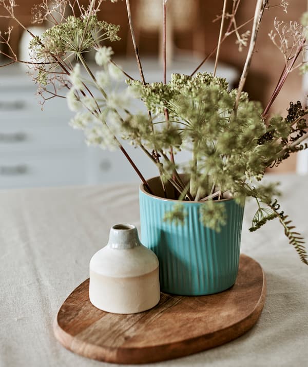 Un cache-pot turquoise utilisé comme un vase avec un bouquet de feuilles sauvages à côté d’un pot en céramique présentés sur une planche à découper.