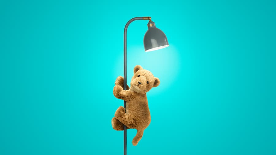 Un cachorro de león de peluche, esponjoso y de color marrón claro, está trepando por una lámpara de pie de color gris oscuro. La escena se sitúa sobre un fondo turquesa vibrante, con una luz brillante que emana de la lámpara.