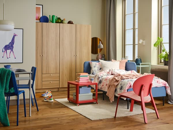 Un cadre de lit avec tête de lit d’angle BLÅKULLEN bleu à côté d’une table d’appoint TINGBY sur roulettes et d’un fauteuil FRÖSET rouge.
