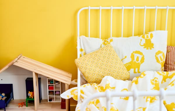 Un cadre de lit blanc avec linge de lit jaune et blanc à motif lion contre un mur jaune, à côté d'une maison de poupée en bois.