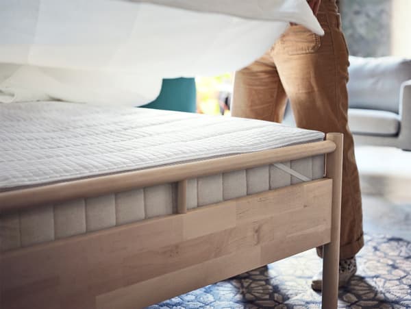 Un cadre de lit en bouleau avec un épais matelas beige et une alèse blanche. Un homme habille son lit de linge de lit blanc.