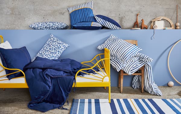 Un cadre de lit jaune avec du linge de lit bleu et blanc avec des imprimés floraux et rayés, des coussins et des jouets en bois sur un mur bleu.