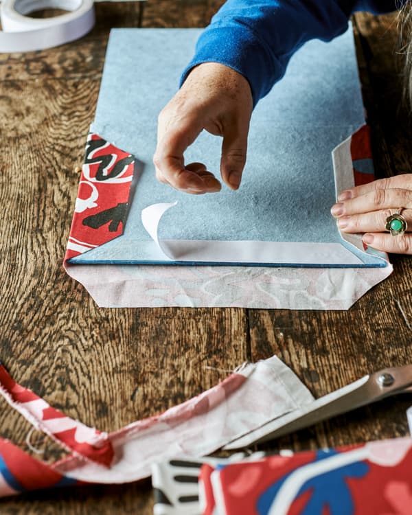 Un cahier bleu ouvert à la page de couverture, des mains collent de l’adhésif double face sur un bord pour y fixer un tissu imprimé rouge.