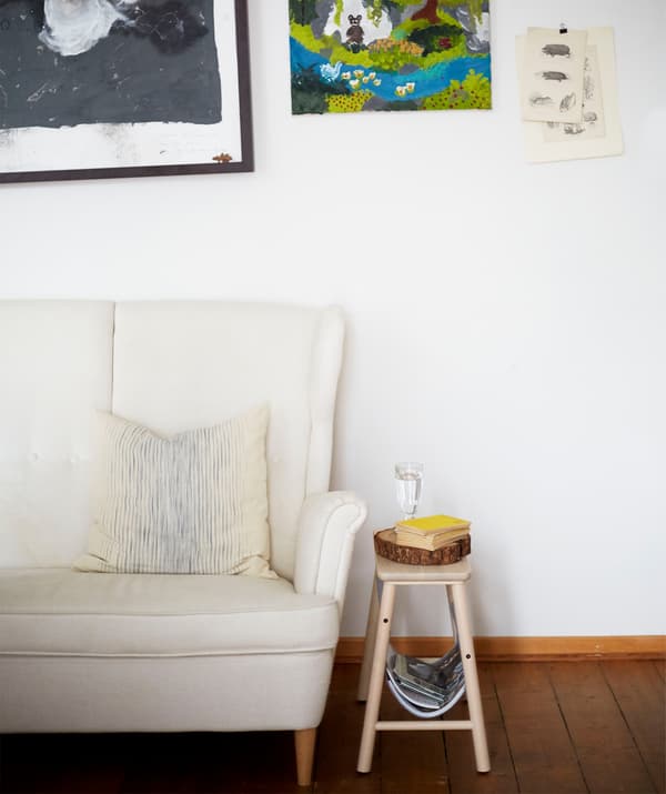 Un canapé blanc à dossier haut et un petit tabouret contre un mur blanc où sont accrochées des œuvres d’art.