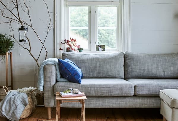 Un canapé gris devant une fenêtre, des plaids en laine rangés dans un panier à côté, et des branches en bois dans un grand vase en verre.