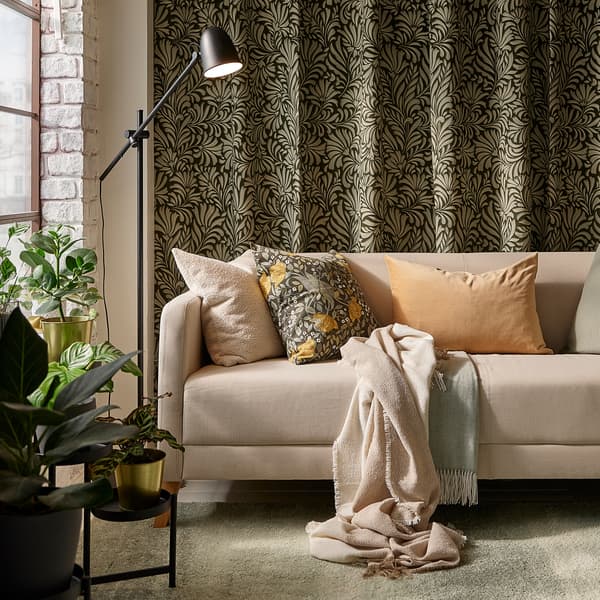 Un canapé LINANÄS beige avec plusieurs coussins sur un tapis gris-vert clair, devant des rideaux à motifs de feuilles accrochés à un mur.