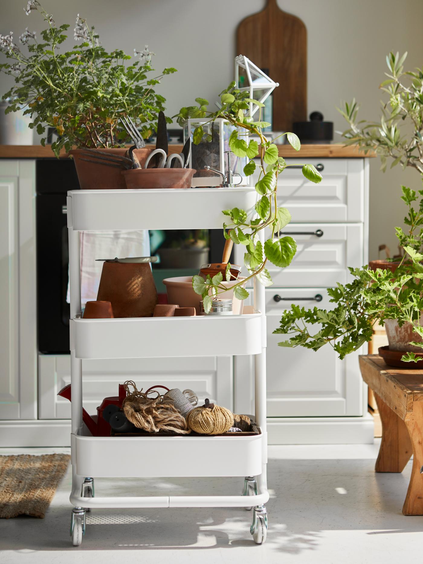 Un carrito RÅSKOG blanco, con macetas, plantas y ovillos de hilo en los estantes, se encuentra en el suelo de una luminosa cocina.
