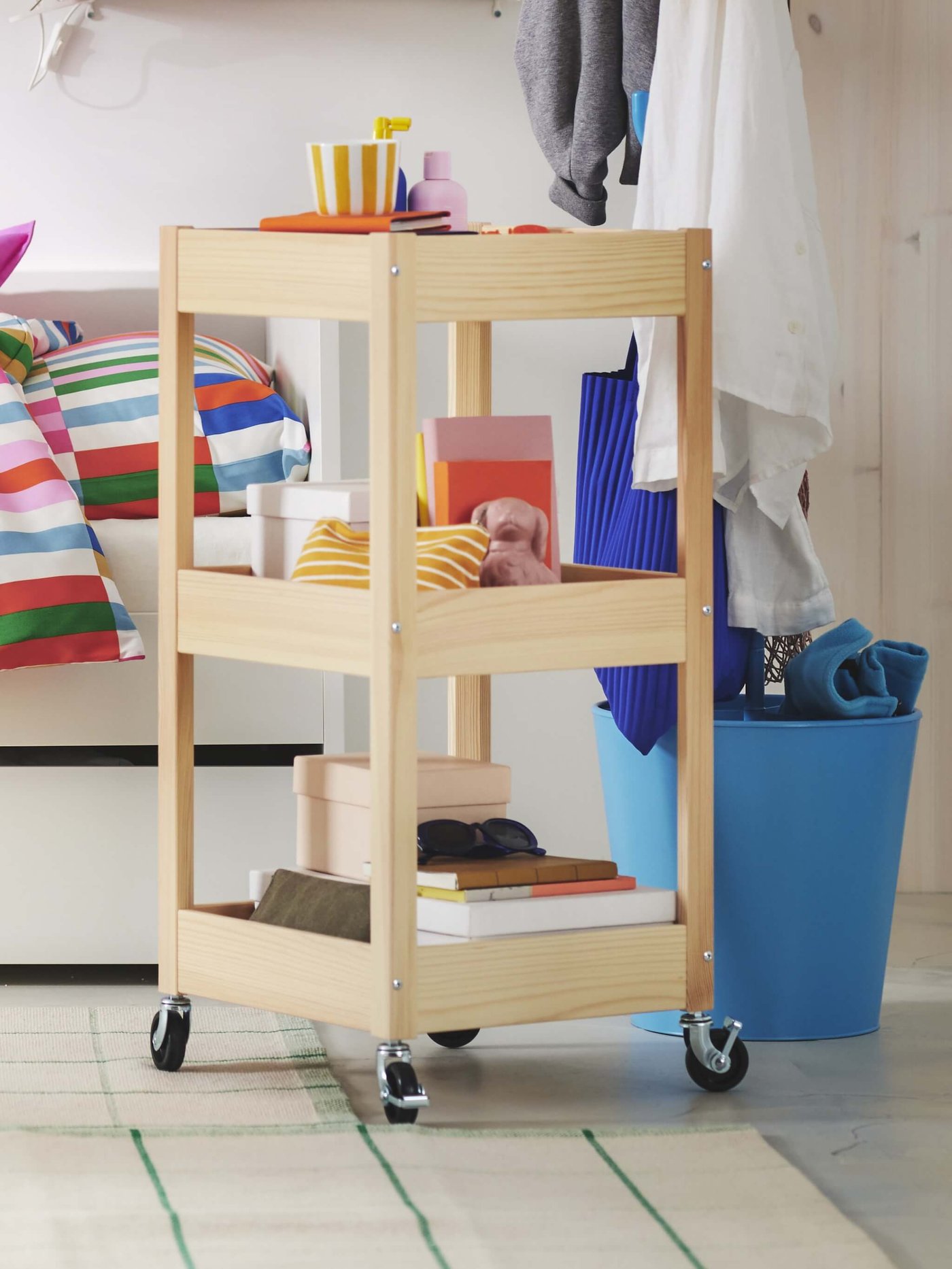 Un carrito rodante de madera con libros, cajas y decoración se encuentra junto a una cama con un edredón colorido, cerca de un contenedor azul con ropa.