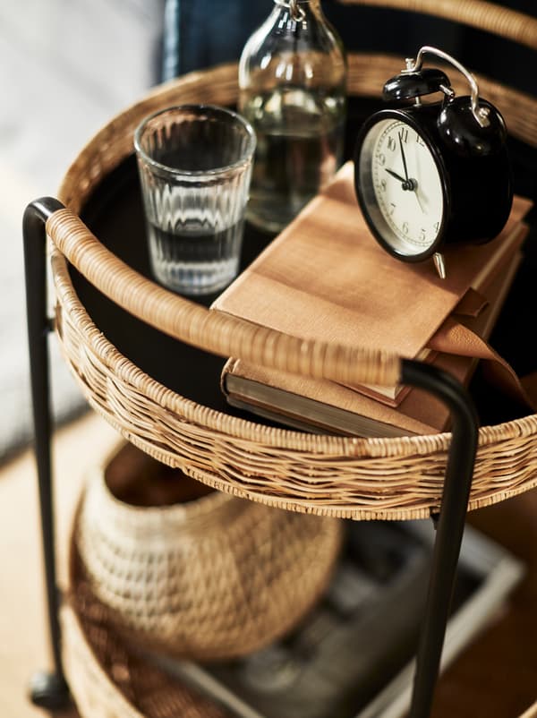 Un cărucior LUBBAN din ratan și metal cu două niveluri, cu o formă rotundă pe care se află cărți, apă și un ceas în partea de sus și diverse obiecte pe polița inferioară.