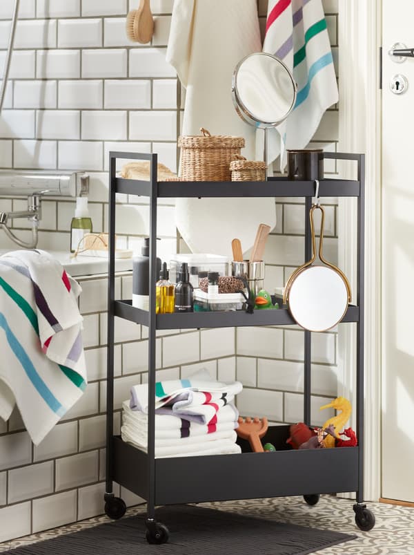 Un cărucior NISSAFORS din oțel negru cu trei polițe pline cu prosoape și jucării de baie este așezat în fața unei căzi.