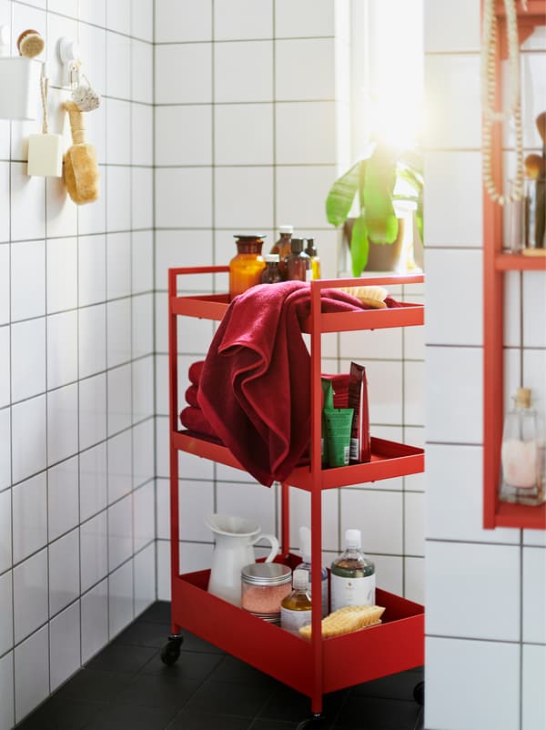 Un cărucior NISSAFORS roșu plin cu accesorii într-o baie cu faianță și gresie albă mobilat tot cu unități ENHET roșii și albe.