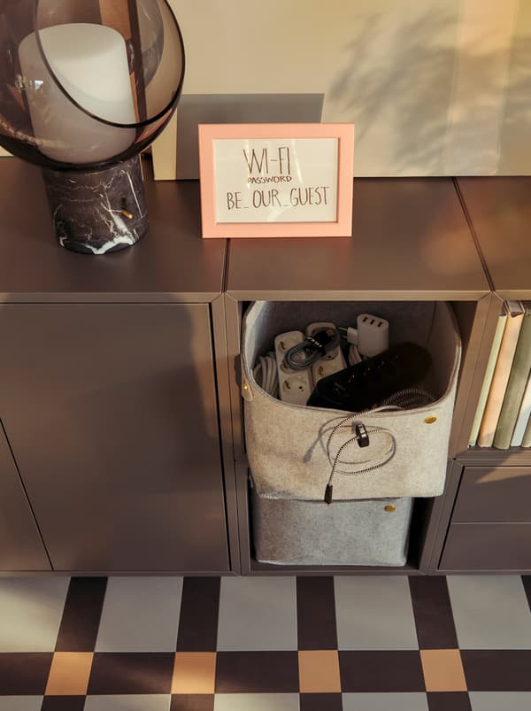 Un cestino PUDDA che contiene accessori elettronici in un mobile basso. La password del Wi-Fi è scritta in un quadretto - IKEA