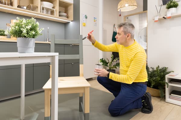 Un co-worker che sta dipingendo una sedia in legno chiaro, verniciandola di colore grigio.