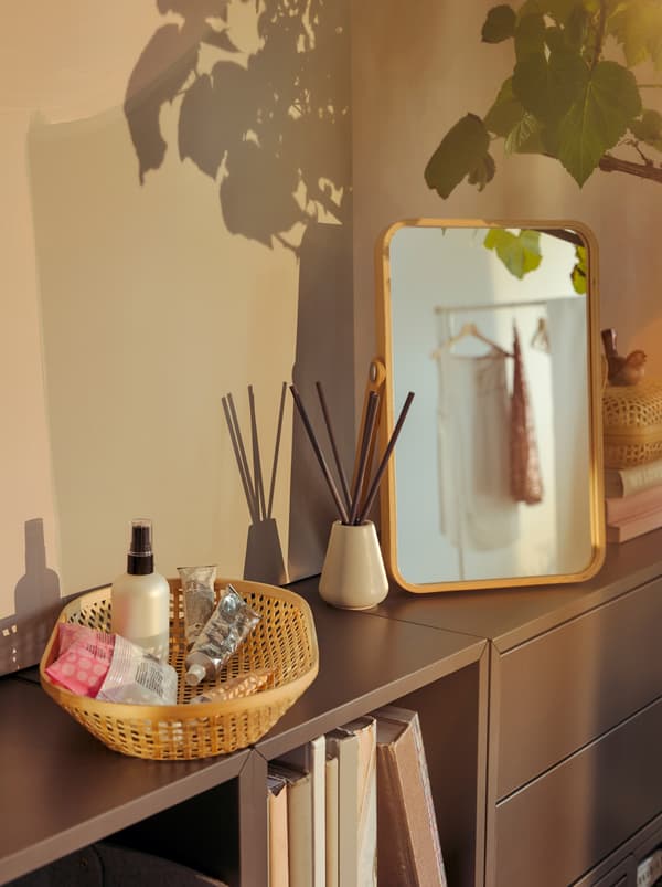 Un coin coiffeuse improvisé sur une bibliothèque basse: des produits de soin dans un panier en bambou KLYFTA, des bâtonnets diffuseurs de parfum NJUTNING et un miroir IKORNNES.
