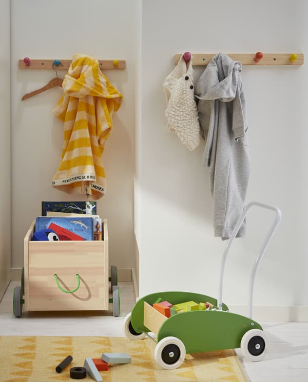 Un coin de jeu avec un chariot de marche vert, un rangement de jouets à roulettes, un tapis jaune, des livres, des jouets et des patères à boutons colorés.