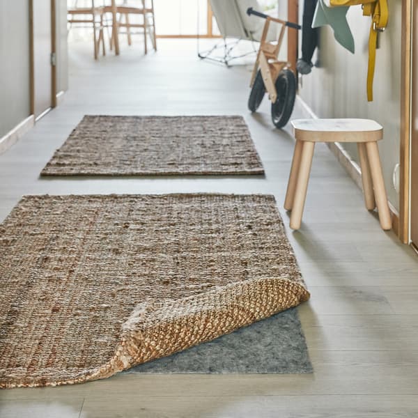 Un coin d’un tapis en jute est relevé et sous celui-ci se trouve une thibaude pour tapis STOPP FILT avec antidérapant qui maintient le tapis en place.