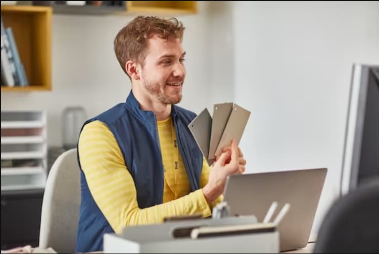 Un colaborador de IKEA sentado en un esritorio sosteniendo tres papeles de tonos grises, mostrándoselos a otra persona.