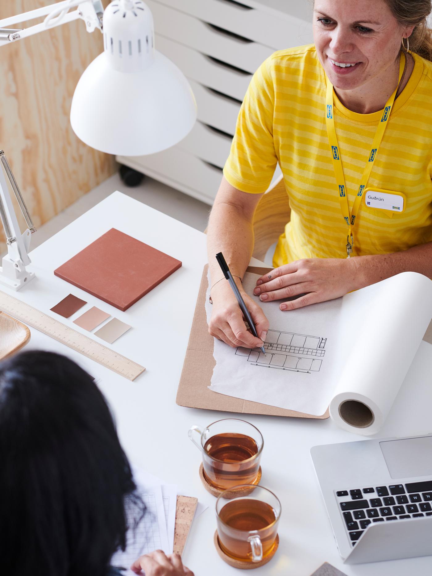 Un collaborateur IKEA à haut jaune s’adresse à un client, prend des notes sur un plan sur un bureau avec une lampe blanche.