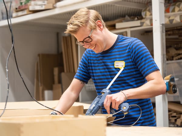 Un collaborateur IKEA portant un uniforme IKEA à rayures bleues sourit en maniant un pistolet à colle électrique dans un atelier.