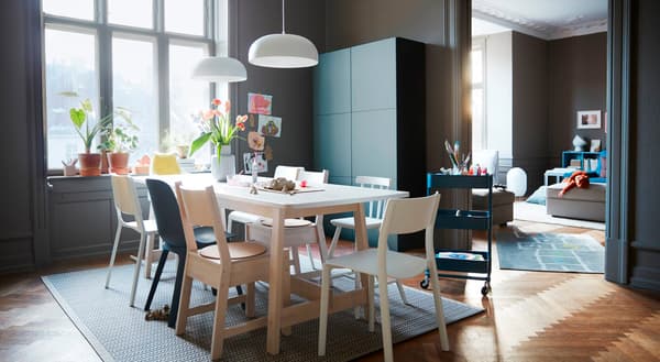 Un comedor con sillas multicolor alrededor de una mesa con la superficie blanca y una combinación de almacenaje gris/turquesa IKEA BESTÅ.