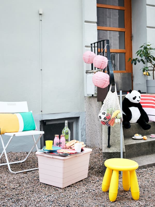 Un contenitore con coperchio SOCKERBIT con sopra bicchieri, posate e ciotole KALAS e una bottiglia KORKEN. Ci sono diversi alimenti e bevande. A lato, uno sgabello giallo MAMMUT, una sieda pieghevole con cuscini. Dietro, un peluche DJUNGELSKOG posato sulle scale.