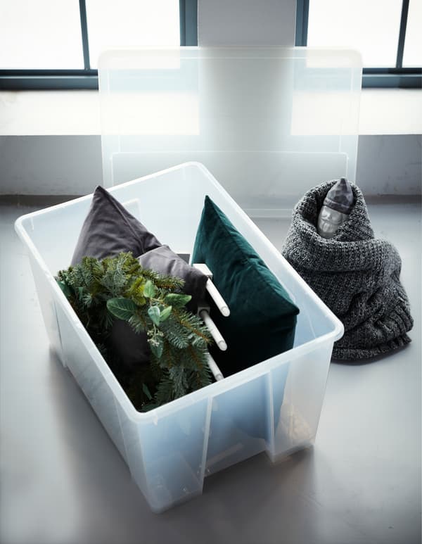 Un contenitore SAMLA in plastica trasparente con le rotelle con dentro due cuscini, una ghirlanda e una lampada. Accanto al contenitore con un nano da giardino avvolto da una coperta.  