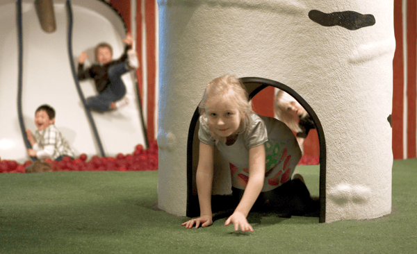 Un copil se târăște printr-un tunel alb cu față de urs; în fundal, doi copii se joacă printre bile roșii pe un covor verde. Fețele sunt blurate.