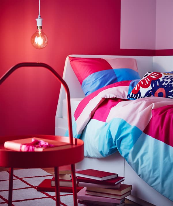 Un copripiumino e una federa multicolore BRUNKRISSLA con fantasia a grandi quadri in diverse tonalità di blu e rosa. In primo piano, un tavolino rosso BURVIK. Sul pavimento, libri impilati. 