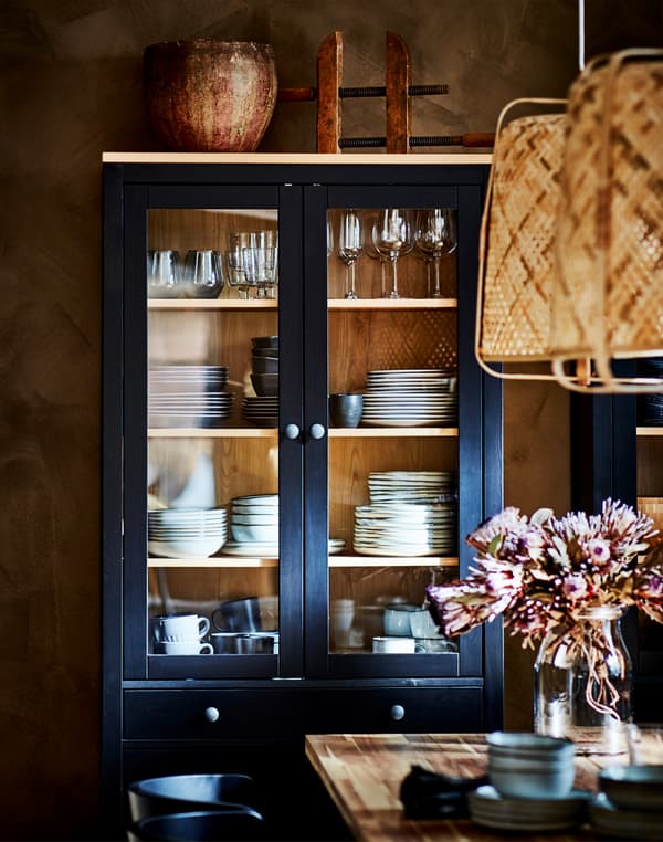 Un corp pentru baie cu uși de sticlă HEMNES în combinația negru/maro unde sunt expuse pahare și veselă, este așezat în spatele unei mese cu o vază de flori.