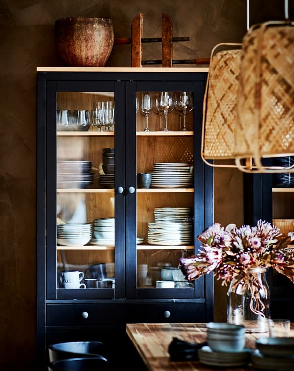 Un corp pentru baie cu uși de sticlă HEMNES în combinația negru/maro unde este expusă veselă este așezat în spatele unei mese cu o vază de flori.