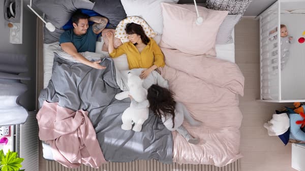 Un couple est allongé dans un grand lit et leur fille joue avec un ourson en peluche. Il y a un bébé à côté dans un lit à barreaux SUNDVIK.