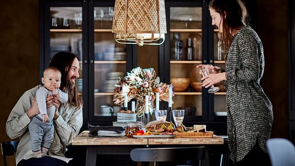 Un couple et un bébé réunis autour d’une table de salle à manger. Vitrines foncées avec des accessoires de table devant le mur du fond.