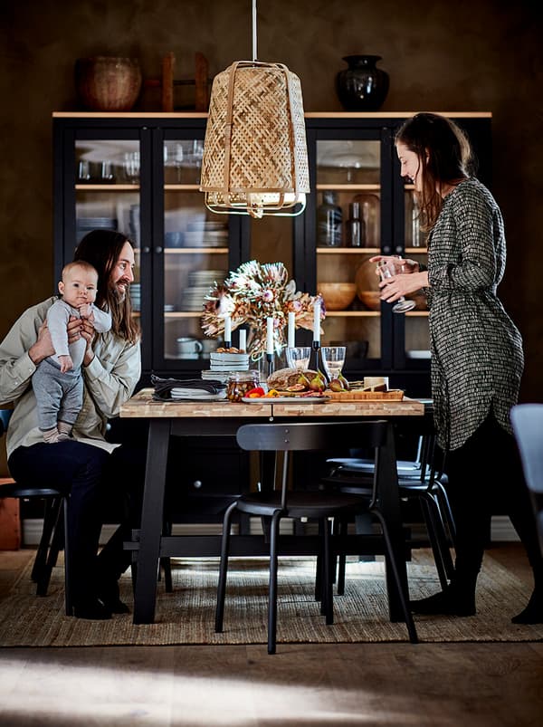 Un couple et un bébé réunis autour d’une table de salle à manger. Vitrines foncées avec des accessoires de table devant le mur du fond.
