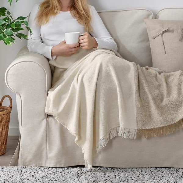 Un coussin dans une housse de coussin AINA beige et un plaid EVALI blanc cassé sur l’accoudoir d’un module d’angle SÖDERHAMN beige clair.