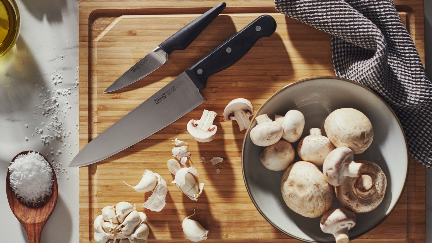Un couteau de chef VARDAGEN, un couteau d’office VÖRDA et de l’ail sur une planche à découper en bambou, en plus d’un bol avec des champignons.