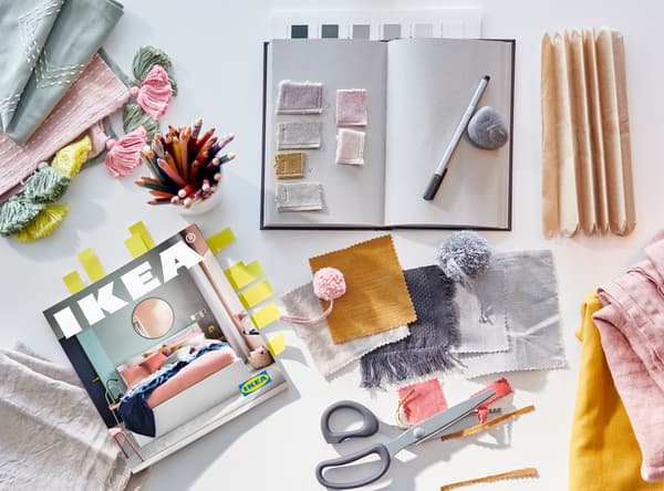 Un cuaderno, un bolígrafo, tijeras y trozos de tela de color rosa, gris, amarillo, mostaza y verde junto a un Catálogo IKEA 2021.