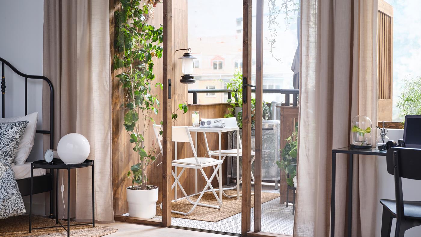 Un cuarto coas portas do balcón abertas no que hai dúas cadeiras e unha mesa FEJAN de cor branca, e moitas plantas.