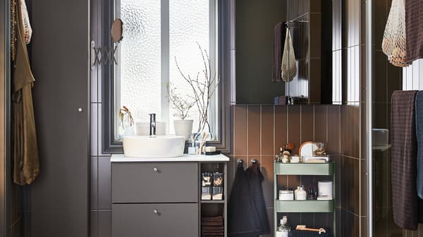Un cuarto de baño con muebles de color gris oscuro, un armario con puerta de espejo y un carrito de color verde grisáceo.