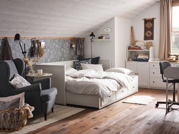 Un divan HEMNES blanc amb tres calaixos en una habitació d'un àtic d'estil rústic amb terra de fusta. Hi ha una butaca STRANDMON al costat.