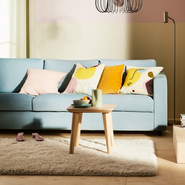 Un divano a 3 posti VIMLE azzurro con diversi cuscini colorati posti sopra e un tappeto VOLLERSLEV a pelo lungo posto di fronte.