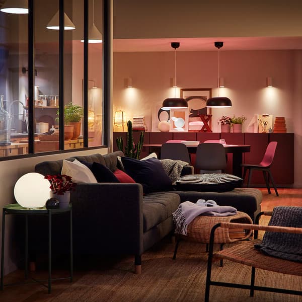 Un divano angolare grigio SOLKLINT con davanti un poggiapiedi contenitore GAMLEHULT in un soggiorno. Sullo sfondo si vede un tavolo ovale con sedie ODGER illuminato da due lampade NYMÅNE.