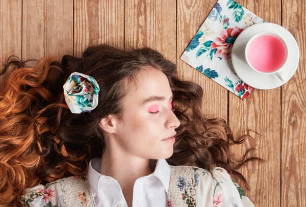 Un donna con una decorazione tra i capelli si riposa su un pavimento in legno. Affianco a lei una tazza di thè con sotto un tovagliolo decorato.