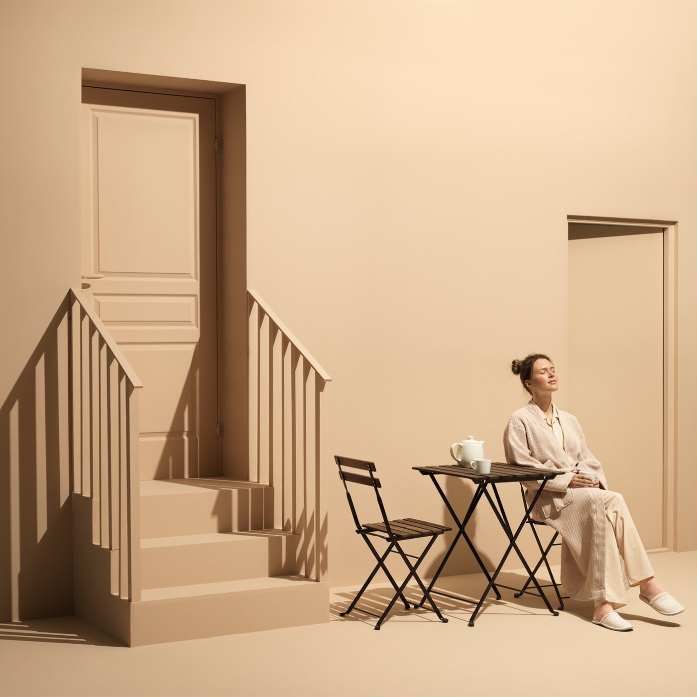 Un donna prende il sole seduta su una sedia TÄRNÖ e con accanto un tavolino e una sedia TÄRNÖ. Le pareti, le porte e le scale sono di color marrone chiaro.